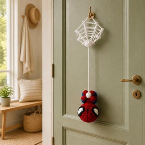 May include: A crocheted wall hanging featuring a white spider web connected by a long cord to a round, red and blue character head with large white eyes. The item is suspended from a gold-coloured metal hook on a sage green panelled door.