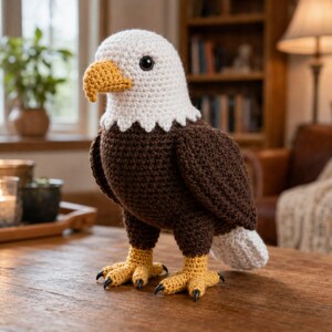 Può includere: Un peluche a forma di aquila calva realizzato all'uncinetto con testa bianca, corpo marrone scuro e coda bianca. L'aquila presenta un becco giallo testurizzato e zampe gialle con artigli neri. Gli occhi sono rotondi, neri e lucidi, posti ai lati.