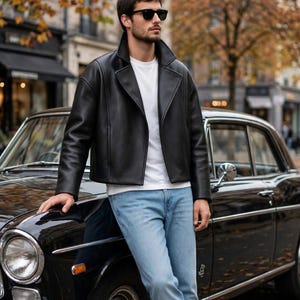 May include: A black leather jacket with a wide collar, a plain white t-shirt, light blue denim jeans, and white sneakers. The outfit is worn with black sunglasses, featuring a smooth texture and a structured fit for a clean, classic appearance.