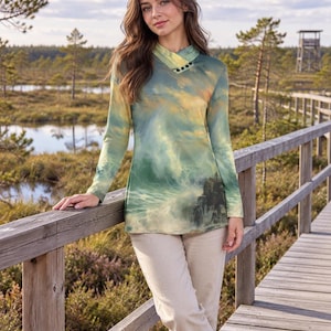 Puede incluir: Una blusa de manga larga con un estampado paisajístico de olas rompiendo y un cielo nublado en tonos verdes, azules y amarillos. La prenda tiene un cuello cruzado adornado con tres pequeños botones negros y se combina con pantalones beige.
