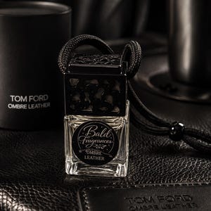 May include: A clear glass car air freshener bottle with a black decorative cap and a black hanging cord. The bottle features a circular label with the text Bald Fragrances and Ombre Leather. A black cylindrical container and a black leather tag are nearby.