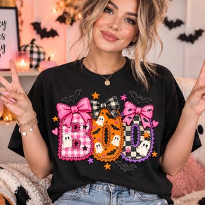 Könnte beinhalten: Ein schwarzes Kurzarm-T-Shirt mit dem Wort BOO in großen Buchstaben. Das B ist rosa kariert mit einer rosa Schleife, das O ist orange mit Fledermäusen und Geistern, und das zweite O ist schwarz-weiß kariert mit einer rosa Schleife und kleinen Geistern.