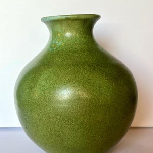 Puede incluir: Un jarrón de cerámica redondo con cuello estrecho y borde acampanado. La superficie presenta un acabado moteado y texturizado en tonos verde hierba y oliva, con sutiles variaciones en la intensidad del color por todo el cuerpo redondeado.