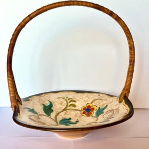 May include: A shallow ceramic bowl with a cream-colored interior featuring a painted floral design in red, yellow, blue, and green. The bowl has a dark brown rim and a tall, arched handle wrapped in light brown wicker material.