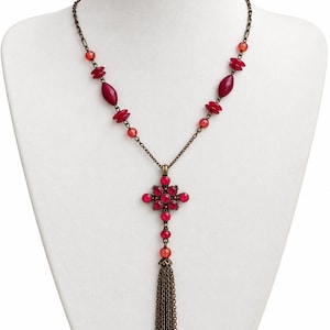 Puede incluir: Un collar con cadena de tono bronce y cuentas rojas y rosa oscuro. El colgante es un diseño en forma de cruz con cristales rojos, suspendido sobre una larga borla de cadenas de bronce que cuelga de una cuenta roja individual.
