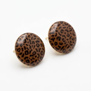 Puede incluir: Un par de pendientes de botón redondos con un patrón de leopardo brillante en marrón y negro. Cada pendiente tiene una superficie lisa y abovedada con un engaste de metal dorado visible en los bordes, diseñado para orejas perforadas.