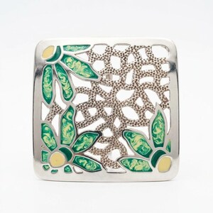 Puede incluir: Un broche cuadrado de metal plateado con un diseño calado y un centro texturizado. La pieza está decorada con motivos florales de esmalte en tonos verde y amarillo pálido, dispuestos en las esquinas del marco cuadrado.