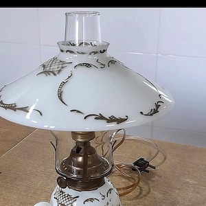 Puede incluir: Una lámpara de mesa con base y pantalla de vidrio opalino blanco decoradas con motivos florales y de celosía en tono dorado. La lámpara incluye un quemador de metal color latón, una chimenea de vidrio transparente y un asa blanca en la base.