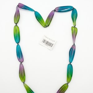 May include: A necklace featuring elongated, smooth beads in a gradient of purple, blue, and green, strung on a thin black cord. A small white rectangular tag with the text MANOUK and a barcode is attached to the center of the necklace.