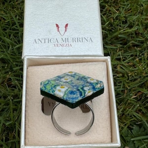 Puede incluir: Un anillo de forma cuadrada con un patrón floral azul y verde con motivos de flores blancas, montado sobre una banda ajustable de tono plateado. El anillo se presenta en una caja blanca con el texto Antica Murrina Venezia y un logotipo rojo.