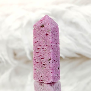 May include: A pink crystal tower with a porous, textured surface featuring small holes and dark mineral inclusions. The stone has a hexagonal prism shape with a pointed top, displaying a vibrant magenta hue throughout its crystalline structure.