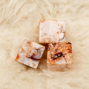 May include: Three polished stone cubes featuring a marbled appearance with a color palette of cream, beige, and burnt orange. Each cube displays unique natural veining and crystalline textures throughout the surfaces, creating a varied pattern.