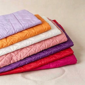 May include: A stack of seven folded fabric pieces featuring a diamond-shaped quilted pattern. The colors include light purple, orange, white, light pink, dark purple, red, and magenta. Each piece has a smooth texture and consistent stitching.