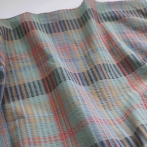 May include: A lightweight fabric featuring a multi-colored plaid pattern with intersecting lines in shades of sage green, muted orange, deep navy blue, and soft yellow. The textile displays a subtle crinkled texture throughout the woven surface.