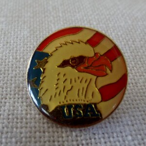 May include: A round pin with a red, white, and blue American flag design. A bald eagle with a yellow beak and red eyes is in the center of the flag. The words "USA" are written in blue below the eagle.
