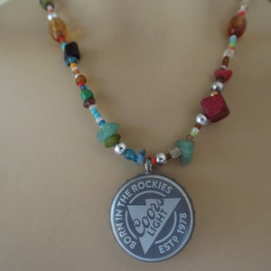 May include: A multi-colored beaded necklace with a silver Coors Light bottle cap pendant. The pendant has the text "Born in the Rockies" and "Est. 1978" on it.