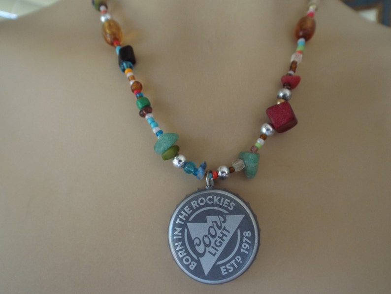 May include: A multi-colored beaded necklace with a silver Coors Light bottle cap pendant. The pendant has the text "Born in the Rockies" and "Est. 1978" on it.