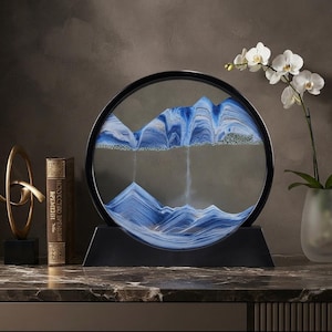 May include: A circular sand art display featuring a black frame and base, containing layers of blue and white sand that form mountain-like shapes. The sand creates a cascading effect within the glass, showcasing distinct peaks and valleys in shades of blue.