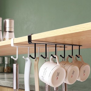 Può includere: Un ripiano portaoggetti in metallo bianco e nero da fissare sotto una mensola, dotato di sei ganci per appendere utensili da cucina. Il supporto è fissato sotto una mensola in legno e sostiene tre tazze in ceramica bianca e vari utensili.