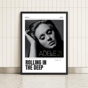 May include: A framed poster featuring a black and white portrait of Adele with her hand near her head. The text reads ADELE, 2010, ADELE 21, ROLLING IN THE DEEP, and 21 ERA / XL. The design is minimalist with a black frame and white border.