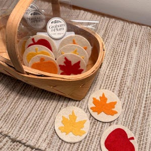 May include: A light wood basket containing several circular felt coasters with autumn-themed designs, including red, orange, and yellow leaves and apples. Each coaster is cream-colored with a felt cutout. A Graham & Plum label is visible on the packaging.