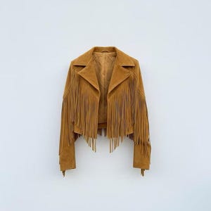 May include: A tan suede jacket featuring long fringe detailing across the front panels and sleeves. The jacket has a wide notched collar, long sleeves, and a smooth interior lining, presented in a cropped length with an open front design.