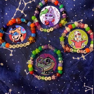 May include: Four colorful beaded bracelets featuring circular character charms. Each bracelet includes letter beads spelling FREDDY, ROXY, CHICA, or MONTY, interspersed with multi-colored plastic beads and star-shaped accents in various bright hues.