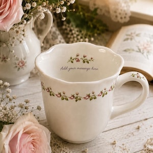 May include: A white ceramic mug with a scalloped rim, featuring a delicate floral garland pattern of small pink roses and green leaves around the exterior and handle. The interior displays the printed text Add your message here.
