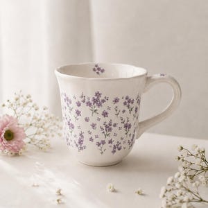May include: A white ceramic mug with a scalloped rim and a handle, featuring a repeating pattern of small purple flowers across the exterior and a single floral detail on the interior rim. The mug has a smooth, glossy finish and a rounded base.