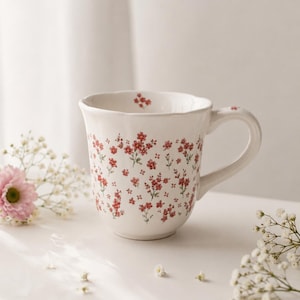May include: A white ceramic mug with a scalloped rim and a handle, featuring a repeating pattern of small red flowers with green stems. A single small red flower is printed on the interior rim, and another is visible on the top of the handle.