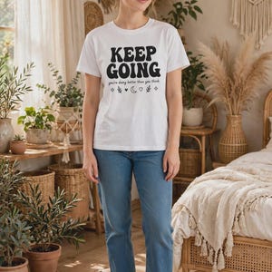 Puede incluir: Una camiseta blanca de manga corta con el texto en negro KEEP GOING, la frase you're doing better than you think y pequeños iconos de estrellas, un corazón y una luna creciente, combinada con vaqueros azules y zapatillas blancas.