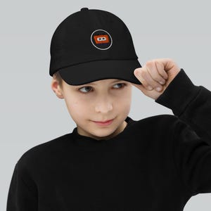 May include: A black baseball cap featuring a circular embroidered patch on the front. The patch displays an orange cassette tape graphic with white details, enclosed within a thin white circular border against the solid black fabric of the cap.