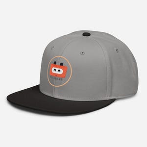 Könnte beinhalten: Eine graue Snapback-Cap mit schwarzem Schirm und schwarzem Knopf oben. Auf der Vorderseite befindet sich ein gesticktes kreisförmiges Logo mit hellorangem Rand, das eine rechteckige orangefarbene Figur mit schwarzen Augen und den Text CODEROBO zeigt.