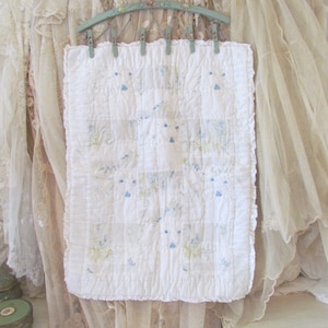 May include: A rectangular white quilted textile featuring a subtle grid pattern and delicate blue floral embroidery. The piece has a ruffled border and is suspended from a light green hanger with small metal clips along the top edge.