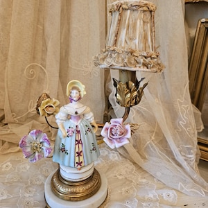 May include: A porcelain figurine lamp featuring a person in a pale blue dress with floral accents, wearing a yellow bonnet. The lamp has a brass base, a fabric shade with lace trim, and decorative ceramic flowers in shades of pink and yellow.
