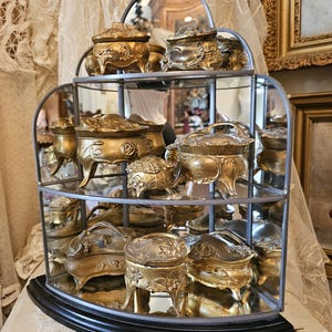 May include: A three-tiered, semi-circular metal display shelf with a mirrored back, holding multiple ornate, gold-toned metal trinket boxes. Each box features intricate embossed floral and leaf patterns, resting on small, curved feet on glass shelves.