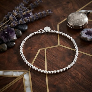 Puede incluir: Una pulsera de cuentas plateadas que presenta una serie de esferas metálicas pulidas y uniformes dispuestas en un círculo. La pulsera incluye una pequeña etiqueta redonda de plata cerca del cierre de mosquetón que une los extremos de la cadena.
