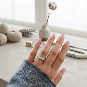 May include: A silver-toned eternity band ring featuring a continuous row of round, clear, faceted stones. The ring is worn on a finger with light pink manicured nails, and the sleeve of a textured, grey, long-sleeved garment is visible at the wrist.