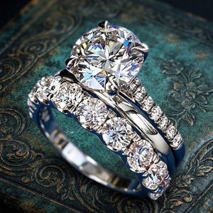 May include: A silver-toned engagement ring set featuring a large round-cut centre stone held by a four-prong setting, accompanied by a matching band. Both rings are adorned with a series of smaller, round-cut stones along the polished metal bands.