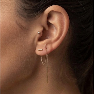 May include: A gold-toned ear piercing set consisting of a small spherical stud in the tragus and a bar stud in the lobe. A delicate gold chain hangs from the bar stud, looping down and back up to create a dangling threader earring effect.
