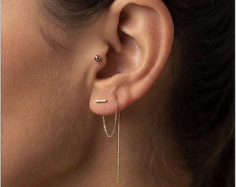 May include: A gold-toned ear piercing set consisting of a small spherical stud in the tragus and a bar stud in the lobe. A delicate gold chain hangs from the bar stud, looping down and back up to create a dangling threader earring effect.