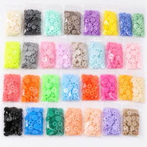 May include: A collection of numerous small plastic snap buttons organized into individual clear plastic bags. The buttons are presented in a wide variety of solid colors, including shades of red, orange, yellow, green, blue, purple, pink, white, black, and tan.