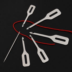 May include: A set of five silver-toned metal latch hook tools with rectangular handles featuring elongated slots, accompanied by a single long, thin sewing needle and a length of red yarn arranged in a curved line across the frame.