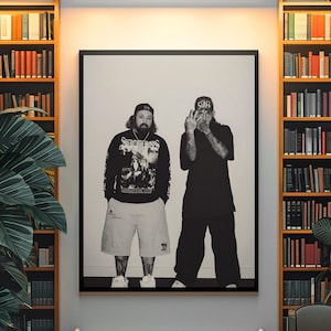 Op de afbeelding: Een zwart-wit ingelijste wandkunstprint met twee personen die naast elkaar staan. De persoon links draagt een shirt met lange mouwen met een afbeelding, een korte broek en een pet. De persoon rechts draagt een t-shirt met het woord STAFF op de pet.