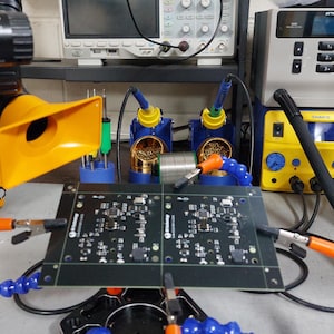 May include: A black printed circuit board held in place by a helping hand tool with blue flexible arms and metal alligator clips. The board features white electronic component markings and is positioned in a workspace with soldering equipment.