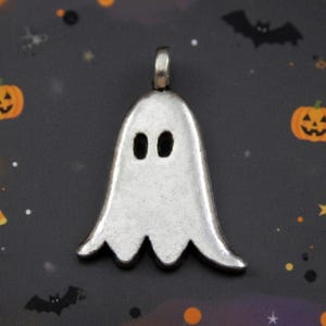 May include: A silver-toned metal pendant shaped like a ghost with two oval-shaped cutouts for eyes. The pendant features a smooth, slightly matte finish and includes a small metal loop at the top for attachment to a necklace or jewelry chain.
