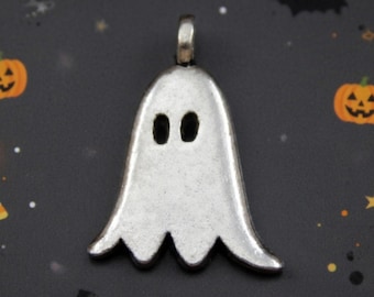 May include: A silver-toned metal pendant shaped like a ghost with two oval-shaped cutouts for eyes. The pendant features a smooth, slightly matte finish and includes a small metal loop at the top for attachment to a necklace or jewelry chain.