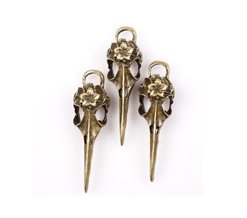 May include: Three gold-toned bird skull charms with floral accents. Each charm features a detailed skull design with a flower on top.