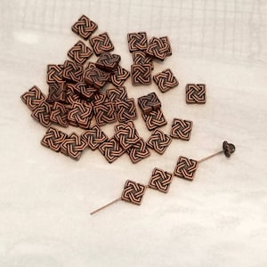 May include: A pile of copper-colored metal square beads with an intricate Celtic knot design. The beads are scattered on a white surface. A few beads are strung on a thin wire.