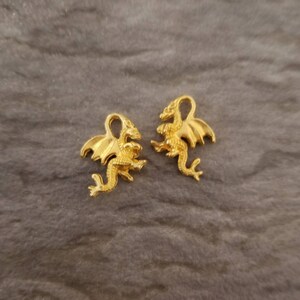 K&ouml;nnte beinhalten: Zwei goldfarbene Drachen-Anh&auml;nger mit Fl&uuml;geln und Schw&auml;nzen. Die Drachen sind in entgegengesetzte Richtungen gerichtet.
